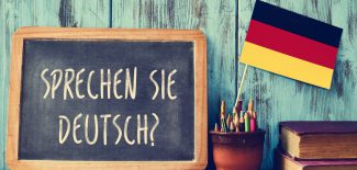 a chalkboard with the question sprechen sie deutsch? do you speak german? written in german, a pot with pencils, some books and the flag of Germany, on a wooden desk